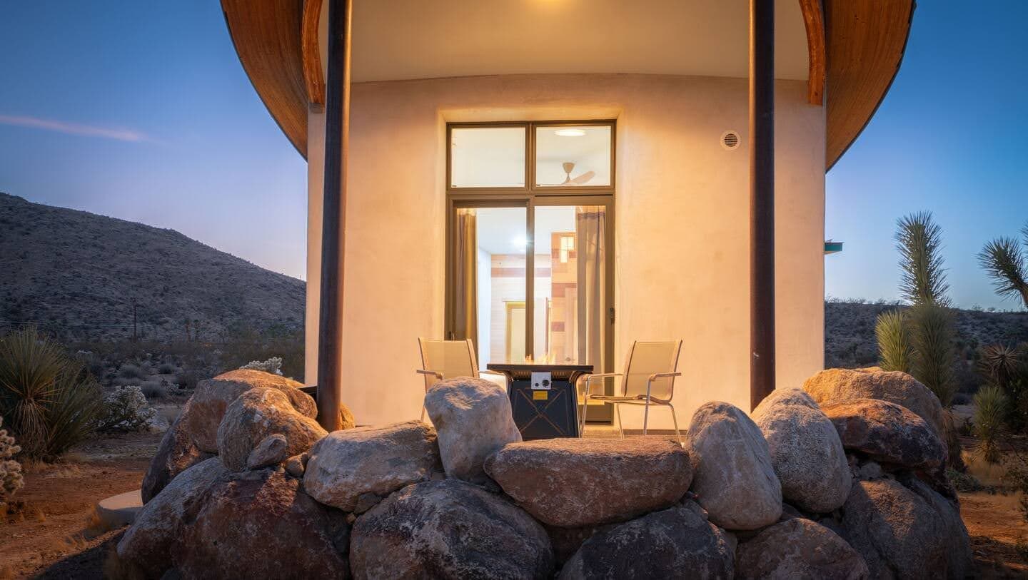 A fire table and seating area on the Mesa Sky Disk patio at dusk, with Joshua trees and boulders around it.